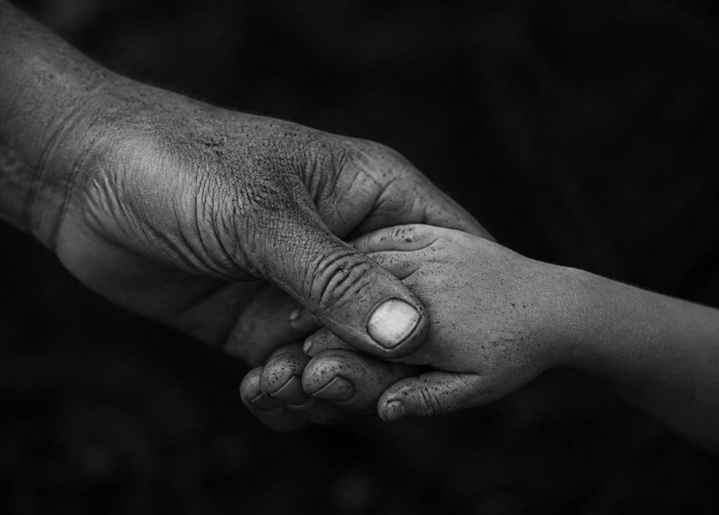 A weathered, soil-covered hand holding a child's hand against a dark background.